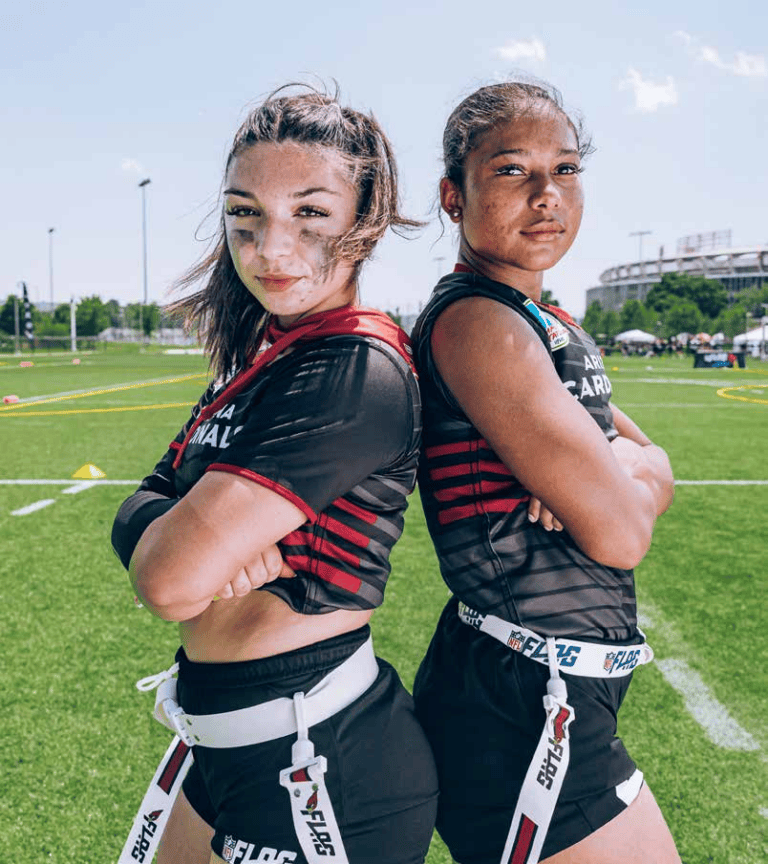 Two students in flag football uniforms stand back to back on the field